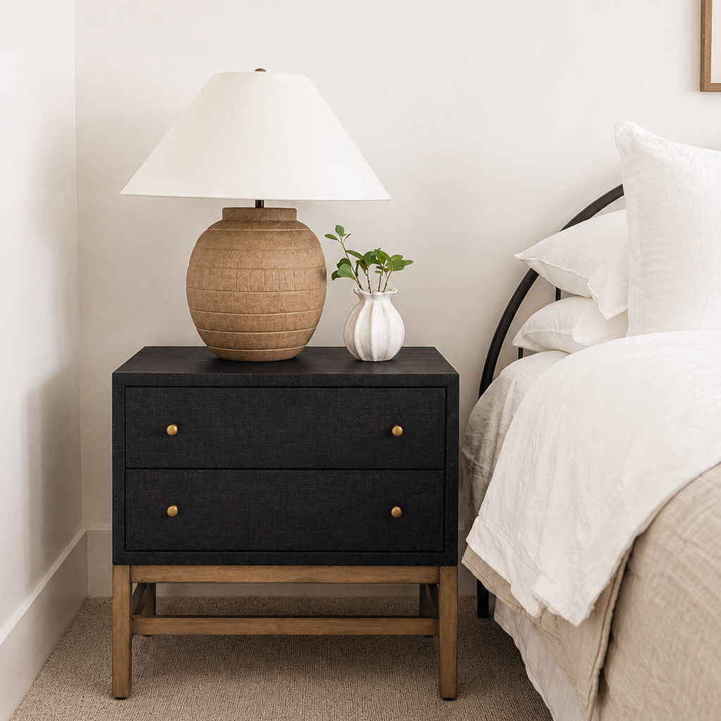 A modern black nightstand with natural wood legs and brass knobs, styled in a bedroom next to a bed with white linens. On top of the nightstand sits a textured beige ceramic table lamp with a white shade and a small white vase with green stems.