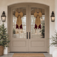 Arched double front doors with glass panes, decorated with two large straw bow ornaments with burgundy ribbon tails, set in a beige entryway with wall sconces, potted plants, and a doormat.